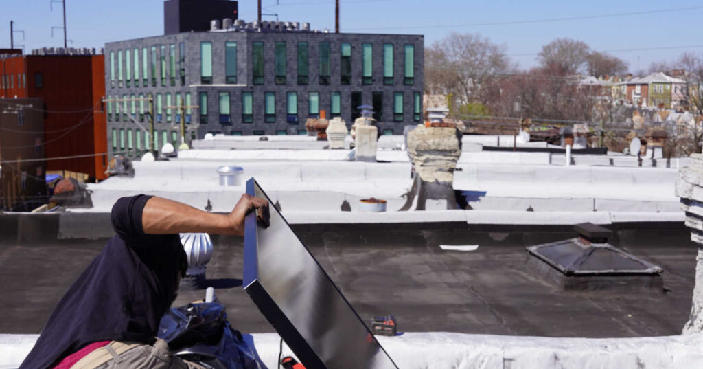 Technician installing a solar panel on a flat roof with city buildings and residential homes visible in the background.