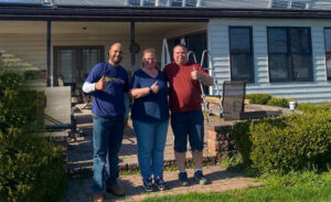 Three people standing in front of a home with rooftop solar panels, giving thumbs up—one wearing a SunWise shirt, indicating a successful solar panel installation.